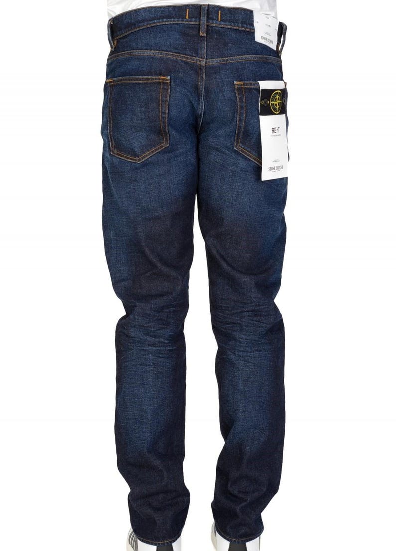 May include: Dark blue denim jeans with a classic five-pocket design. The jeans have a straight leg fit and a faded appearance. A white tag with black text is attached to the back pocket. The jeans are worn with white sneakers.