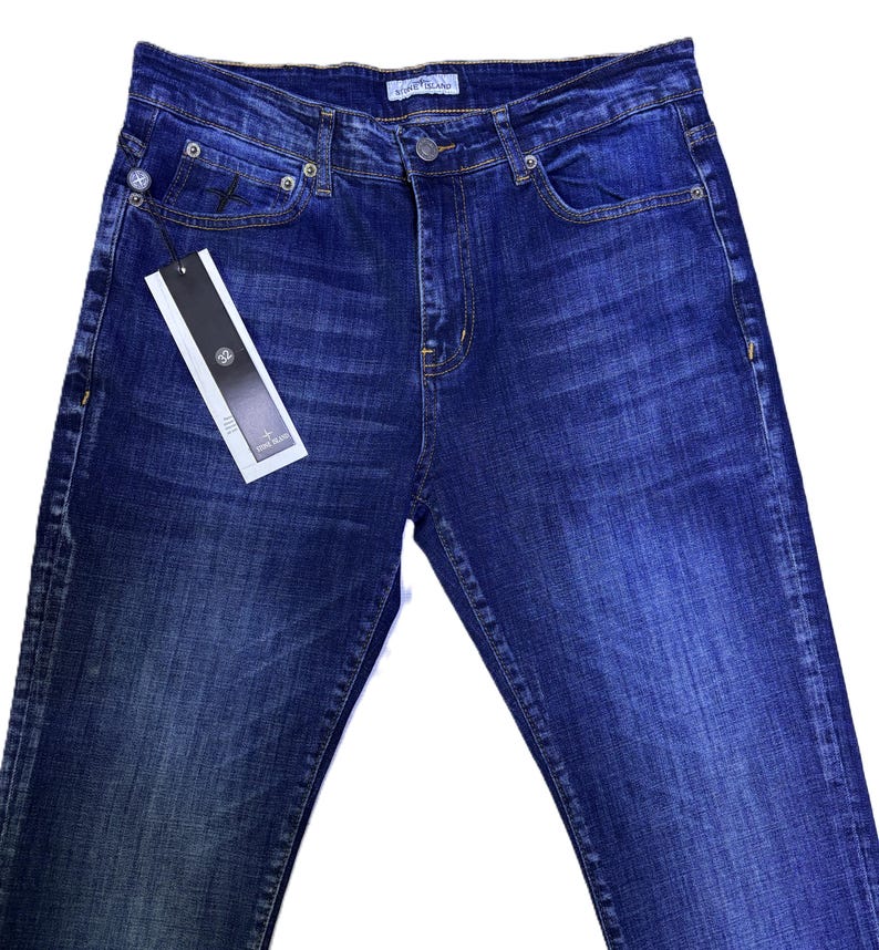 May include: Dark blue denim jeans with a classic five-pocket design. The jeans have a button and zipper closure, belt loops, and a small tag attached to the front pocket. The denim has a faded wash effect.