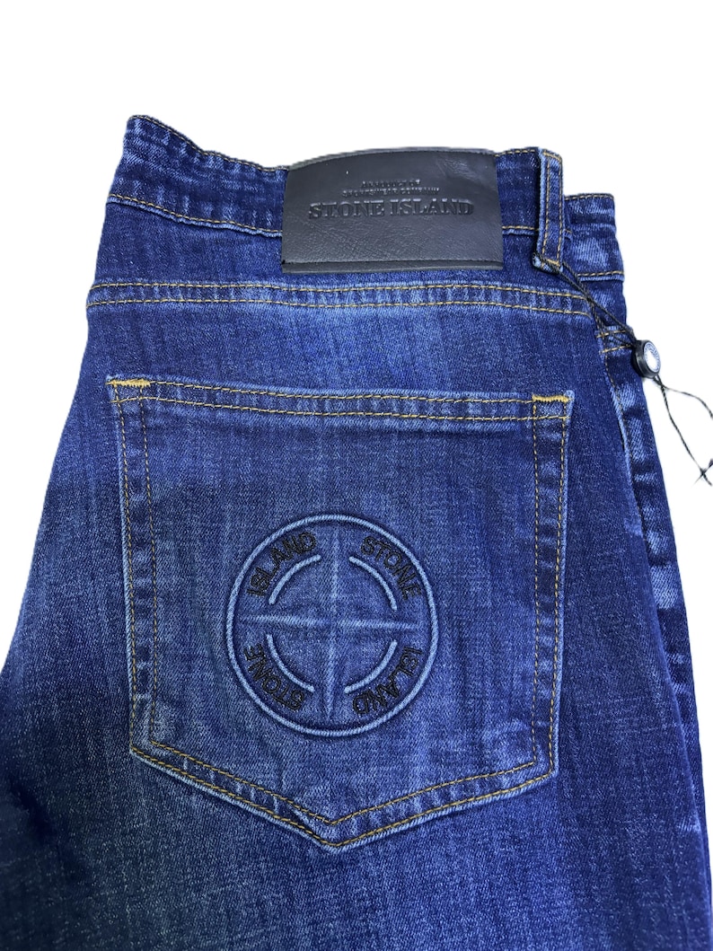 May include: Dark blue denim jeans with a classic five-pocket design. Features a leather patch with "STONE ISLAND" branding and a circular logo on the back pocket. The jeans have a straight leg cut and a button and zipper closure.