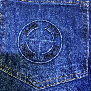 May include: Close-up of a blue denim pocket with a stitched circular logo. The logo features the words "STONE ISLAND" and a compass rose design. The pocket has yellow stitching.