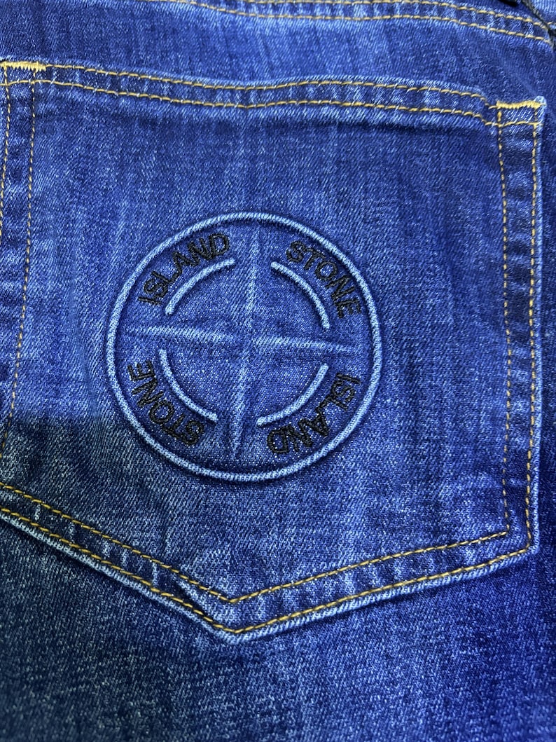 May include: Close-up of a blue denim pocket with a stitched circular logo. The logo features the words "STONE ISLAND" and a compass rose design. The pocket has yellow stitching.