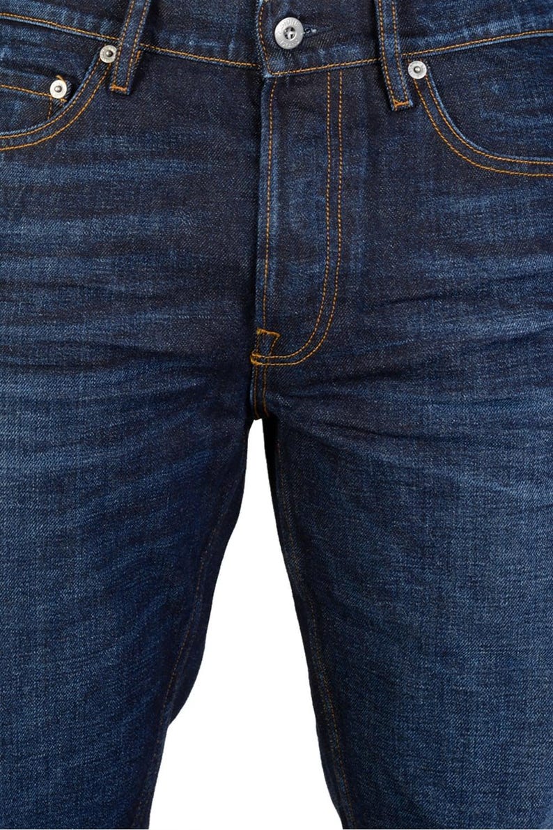 May include: Close-up of dark blue denim jeans. The jeans feature classic five-pocket styling with orange stitching. The fly has a button closure, and the rivets and button are silver. The fabric has a textured appearance.