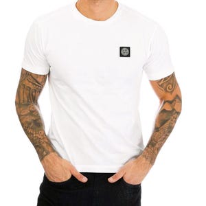Può includere: T-shirt bianco girocollo con un piccolo logo quadrato sul petto. La maglietta a maniche corte ha una vestibilità classica. La persona che indossa la maglietta ha tatuaggi su entrambe le braccia.