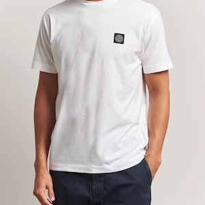 Peut inclure: Un t-shirt blanc à manches courtes avec un petit écusson carré sur le côté gauche de la poitrine. L'écusson est noir avec un emblème circulaire au centre. La personne porte un pantalon bleu marine. Le t-shirt est en matière douce et confortable.