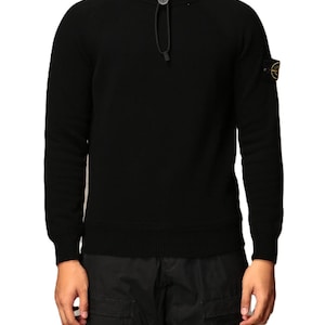 Può includere: Maglione nero con cappuccio, collo alto e chiusura con coulisse. Il maglione presenta una piccola toppa con logo sulla manica sinistra. Le maniche sono lunghe e il materiale sembra essere un tessuto a maglia. Il maglione è indossato con pantaloni cargo neri.