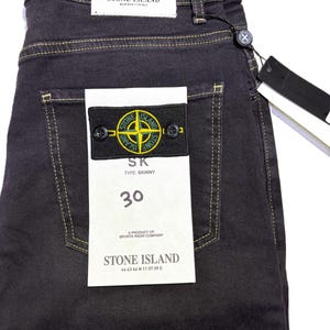 May include: Dark gray denim jeans with yellow stitching and a Stone Island patch on the back pocket. The jeans have a size 30 label and a black tag. The Stone Island logo is a compass rose.