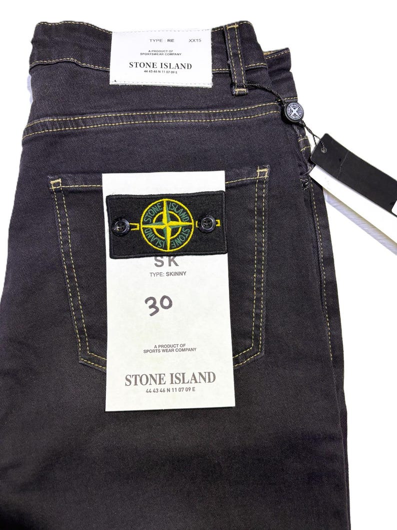 May include: Dark gray denim jeans with yellow stitching and a Stone Island patch on the back pocket. The jeans have a size 30 label and a black tag. The Stone Island logo is a compass rose.