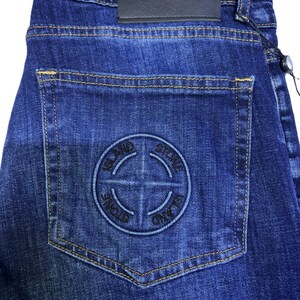 May include: Dark blue denim jeans with a stitched logo on the back pocket. The jeans have a leather patch with the brand name and a tag attached. The stitching is a contrasting yellow color.