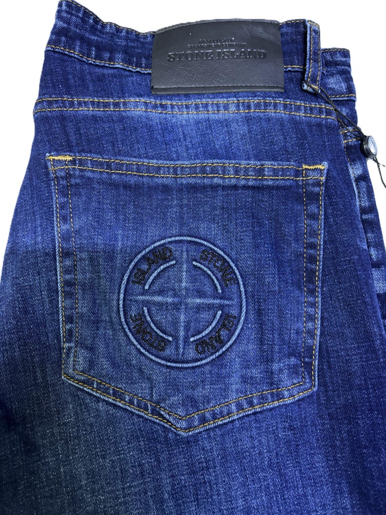 May include: Dark blue denim jeans with a stitched logo on the back pocket. The jeans have a leather patch with the brand name and a tag attached. The stitching is a contrasting yellow color.
