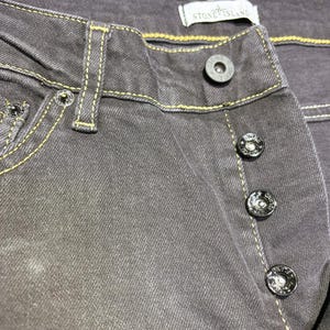 May include: Close-up of gray denim jeans with yellow stitching. The jeans have a button fly and a Stone Island label. The buttons are black with a silver rim and the Stone Island logo.