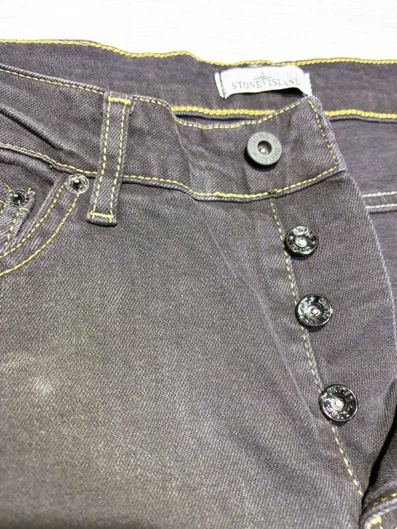 May include: Close-up of gray denim jeans with yellow stitching. The jeans have a button fly and a Stone Island label. The buttons are black with a silver rim and the Stone Island logo.
