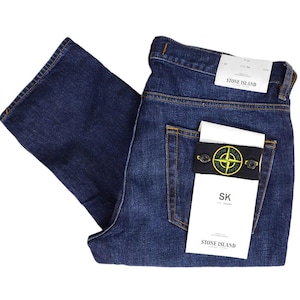 May include: Dark blue denim jeans with a classic five-pocket design. The jeans feature a Stone Island label on the back pocket, with a black and yellow logo patch. The jeans are folded, showcasing the back pockets and the brand's tag.