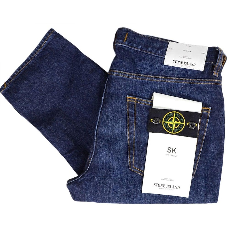 May include: Dark blue denim jeans with a classic five-pocket design. The jeans feature a Stone Island label on the back pocket, with a black and yellow logo patch. The jeans are folded, showcasing the back pockets and the brand's tag.