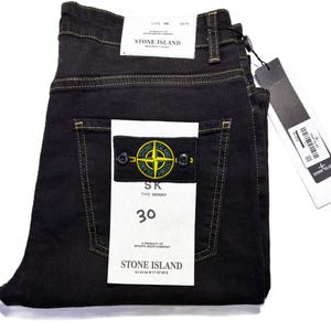 May include: Black skinny jeans with yellow stitching, featuring a Stone Island patch on the back pocket. The jeans have a size 30 label and a Stone Island tag. The jeans are folded neatly.