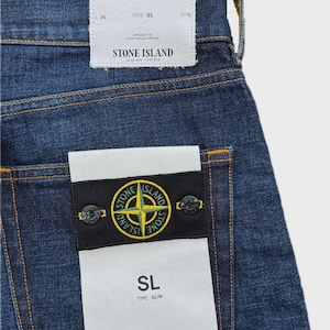 May include: Close-up of blue denim jeans with a white tag that reads "STONE ISLAND". The jeans have a black patch with a yellow and green logo. The jeans are size W36.