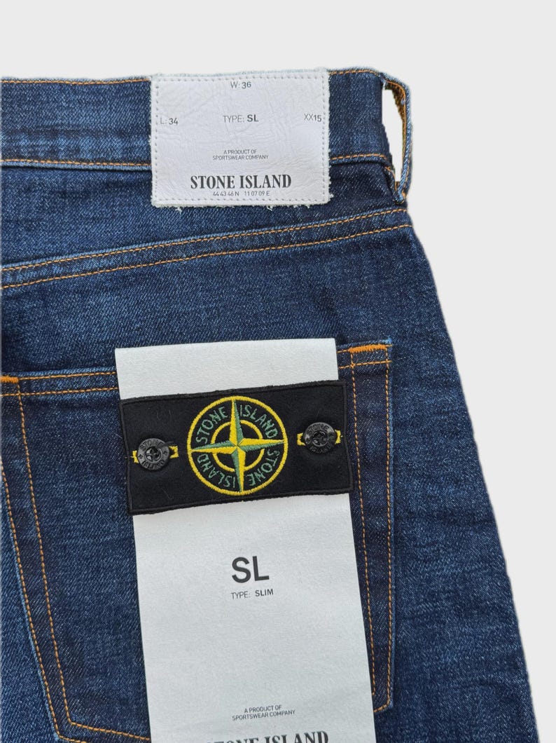 May include: Close-up of blue denim jeans with a white tag that reads "STONE ISLAND". The jeans have a black patch with a yellow and green logo. The jeans are size W36.