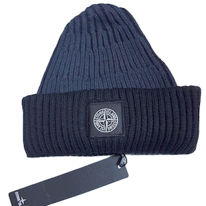 Puede incluir: Un gorro de punto acanalado azul marino y negro. El gorro presenta un parche cuadrado con un logotipo de rosa de los vientos. Una etiqueta negra está adherida al gorro.