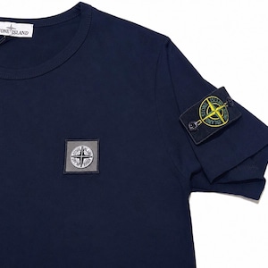 Puede incluir: Camiseta azul marino con cuello redondo y mangas cortas. Presenta un pequeño parche cuadrado en el pecho y otro más grande en la manga izquierda, ambos con un diseño de logotipo. La marca "STONE ISLAND" está impresa en una etiqueta.