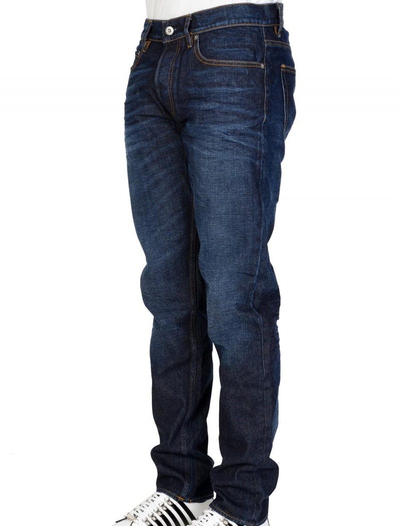 May include: Dark wash denim jeans with a classic five-pocket design. The jeans have a straight leg fit and are worn with white sneakers. The denim has a slight fade and subtle texture.