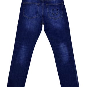 May include: Dark wash denim jeans with a classic five-pocket design. Features a black leather patch on the back waistband and a circular emblem on the back right pocket. The jeans have a straight leg cut.