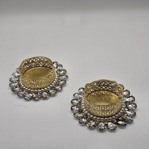 May include: Two decorative candle holders. Each has a gold-colored, openwork metal cup set on a base of clear, faceted crystals and small white pearls. The candle holders are on a white surface.