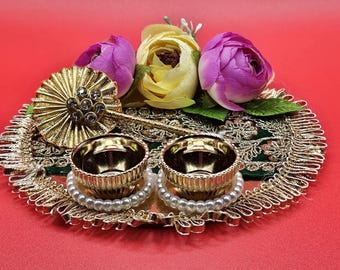 May include: A decorative gold tray featuring two small gold bowls, edged with a pearl trim. The tray is decorated with artificial purple and yellow flowers, and a gold embellishment. The background is red.