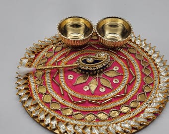 May include: A decorative pink and gold plate with two small gold bowls. The plate features intricate gold and silver embellishments, with a central design and a small spoon-like object. A cotton bud rests on the plate.