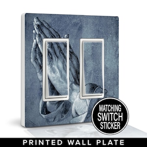 May include: Printed wall plate featuring a detailed illustration of praying hands in shades of blue and gray. The wall plate has two light switch openings with white frames. A matching switch sticker is included.