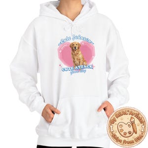 Puede incluir: Sudadera con capucha blanca con bolsillo delantero y cordón. La parte delantera presenta un gráfico rosa y azul con un golden retriever y el texto "Cute Johnson" y "CUTE ATTACK Good day".