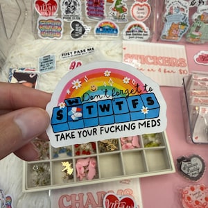 Peut inclure: Un autocollant découpé représentant un arc-en-ciel au-dessus d'un pilulier bleu avec les lettres S, M, T, W, T, F, S. Le texte indique Don't forget to TAKE YOUR FUCKING MEDS. Le motif inclut de petites fleurs blanches et un smiley.