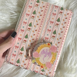 May include: A clear phone case with a pink Christmas pattern featuring Santa, Christmas trees, and bows. A decorative pop socket with a purple dragon design and pink roses is attached to the case.