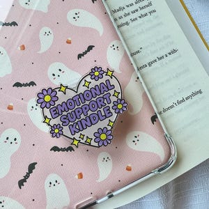May include: A pink phone case with a repeating pattern of white ghosts, black bats, and orange candy corn. A heart-shaped sticker with the text "EMOTIONAL SUPPORT KINDLE" is on the case, surrounded by purple flowers and yellow stars.