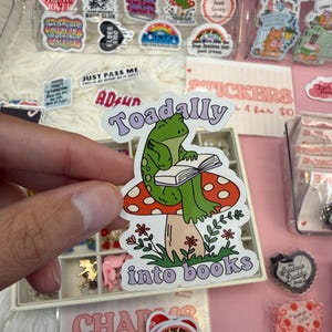 Puede incluir: Una pegatina de vinilo troquelada que muestra una rana verde sentada sobre una seta roja con puntos blancos mientras lee un libro abierto. El texto "Toadally" aparece en morado sobre la rana y "into books" está escrito en morado bajo la seta.