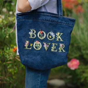 May include: A dark blue denim tote bag featuring the embroidered text BOOK LOVER in light green block letters. Each letter is intricately decorated with colorful floral motifs, including small red, yellow, and blue flowers and green leaves.