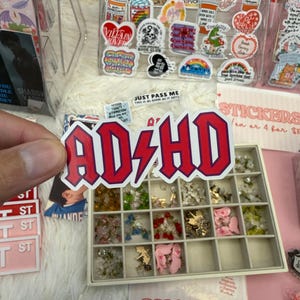 Op de afbeelding: Een gestanste vinylsticker met de tekst AD/HD in een vetgedrukt, rood gotisch lettertype met een paarse omlijning. De letters zijn in het midden verbonden door een rood bliksemschichtsymbool, alles op een strakke witte achtergrond.