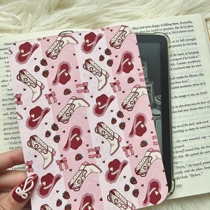 Może przedstawiać: Różowa i biała wzorzysta okładka na Kindle z kowbojskimi butami, kapeluszami, truskawkami i kokardkami. Okładka jest otwarta, ukazując ekran czytnika i książkę.