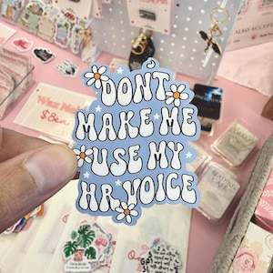 Può includere: Un adesivo azzurro fustellato con il testo bianco "DONT MAKE ME USE MY HR VOICE" in un carattere ondulato di ispirazione retrò. Il design è decorato con piccole stelle bianche e margherite bianche con il centro arancione.