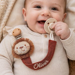 Peut inclure: Une attache-sucette avec une tête de lion en peluche dotée d'une crinière à franges marron, fixée à une sangle en tissu rouille brodée du prénom Oliver en écriture blanche, reliée à une pince métallique avec un bouton en bois rond.