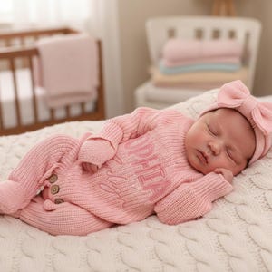 Op de afbeelding: Een pasgeboren baby draagt een roze gebreide outfit en bijpassende hoofdband met strik, liggend op een witte kabelgebreide deken. De outfit is geborduurd met de naam "Sophia". Een wieg en andere babyartikelen zijn op de achtergrond te zien.