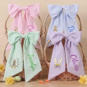 Puede incluir: Cuatro lazos de tela a rayas en rosa, verde, azul y morado, cada uno con un nombre bordado en las colas. Los lazos están en cestas de mimbre, con bordados y diseños temáticos de Pascua. Decoración de Pascua en primer plano.