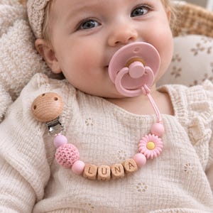 May include: A pacifier clip featuring a round wooden clip, a pink crocheted bead, smooth pink silicone beads, a pink daisy-shaped bead, and wooden letter beads spelling the name LUNA. The clip is attached to a light pink silicone pacifier.