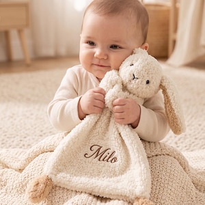 May include: A cream-colored plush security blanket featuring a stuffed bunny head with long ears and brown embroidered details. The soft, textured fabric includes the name Milo stitched in brown script lettering on the front corner.