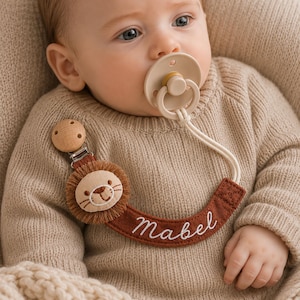 Puede incluir: Un chupete beige unido a un cordón color crema y una cinta de tela color óxido con el nombre Mabel bordado. La cinta incluye una cabeza de león de peluche con melena de flecos marrones y un clip de madera redondo con cierre metálico.