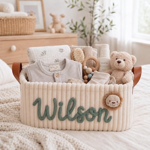 May include: A cream-colored corduroy storage basket with brown leather handles, featuring the name Wilson in green cursive yarn lettering and a small lion face patch. The basket contains a baby bodysuit, a muslin cloth, a wooden rattle, and a teddy bear.