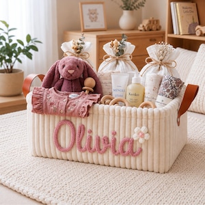 Può includere: Un cesto portaoggetti in tessuto a coste color crema con manici in pelle marrone e il nome Olivia ricamato in filo rosa sul davanti, contenente un coniglio di peluche viola, una camicia rosa con volant, prodotti per la cura e giocattoli in legno.