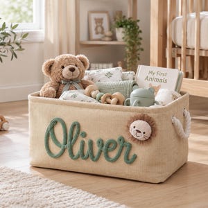 May include: A rectangular beige fabric storage basket featuring the name Oliver in raised green script lettering and a small plush lion face appliqué. The basket includes white rope handles and is filled with a teddy bear, a book titled My First Animals, and various baby items.