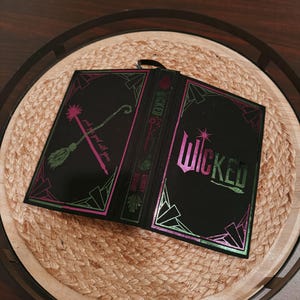 May include: Black book-shaped box with the word "Wicked" in green and pink lettering. The box has a decorative border and features a broom and text that reads "pink is good with green". The box is open and sits on a woven tray.
