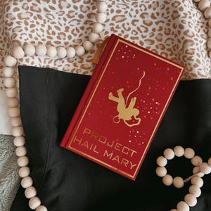 Op de afbeelding: Een rood boek met de titel "PROJECT HAIL MARY" met een gouden astronautenillustratie. Het boek heeft een gouden bladwijzer en ligt op een zwart oppervlak. Een houten kralenkrans en een patroonstof bevinden zich op de achtergrond.