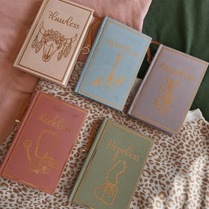 Könnte beinhalten: Fünf Hardcover-Journale in Pastellfarben. Jedes Journal hat goldfarbenen Text und Illustrationen, darunter ein Bullenschädel, eine Gitarre, Ballettschuhe, ein Stethoskop und Hundemarken. Mit einem Leder-Lesezeichen.