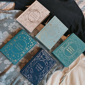 May include: A collection of hardcover books with decorative covers. The books have titles like "A Court of Thorns and Roses" and "A Court of Silver Flames." The covers are in various colors, including blue, teal, and cream, with gold and silver lettering.